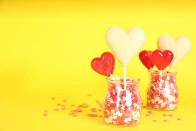 Photo of Heart shaped lollipops made of chocolate and sugar syrup with sprinkles in glass jars on yellow background. Space for text