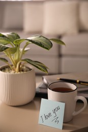 Photo of Paper note with handwritten words I Love You, cup of tea and houseplant on table indoors