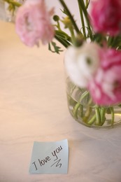 Photo of Paper note with handwritten text I Love you on white table near flowers. Romantic message