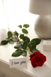 Photo of Paper with text I Love You and red rose on nightstand indoors