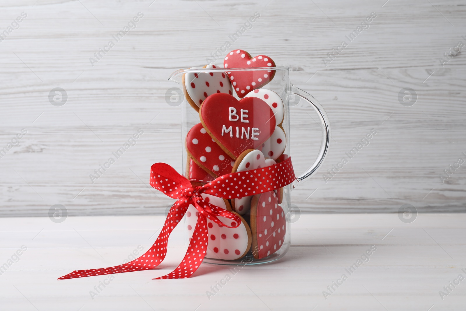 Photo of Delicious heart shaped cookies in glass jug with red bow on white wooden table