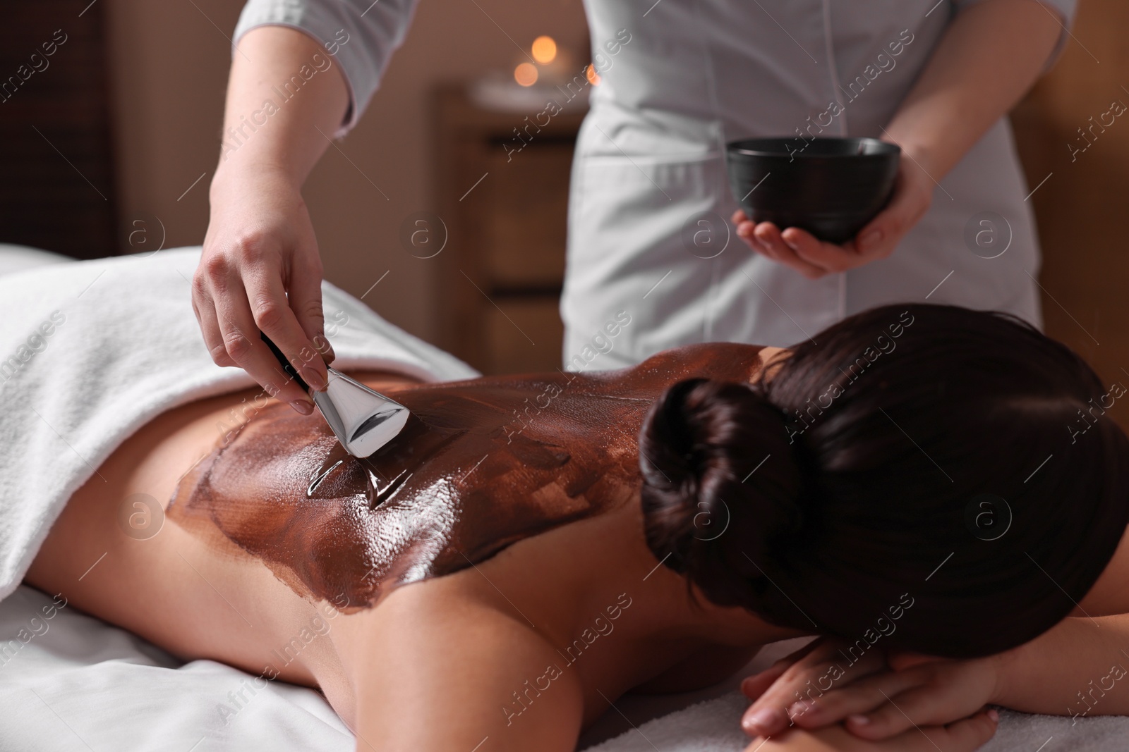 Photo of Chocolate body wrap. Spa worker applying mask onto woman's back in salon, closeup