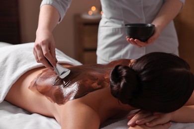 Photo of Chocolate body wrap. Spa worker applying mask onto woman's back in salon, closeup