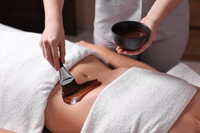 Photo of Chocolate body wrap. Spa worker applying mask onto woman's belly in salon, closeup