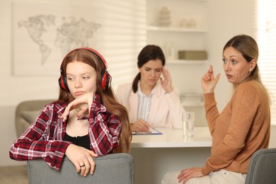 Photo of Annoyed teenage girl listening to music instead of working with psychologist in office, selective focus. Mother and daughter difficult relationship