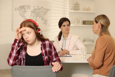 Photo of Annoyed teenage girl listening to music instead of working with psychologist in office, selective focus. Mother and daughter difficult relationship