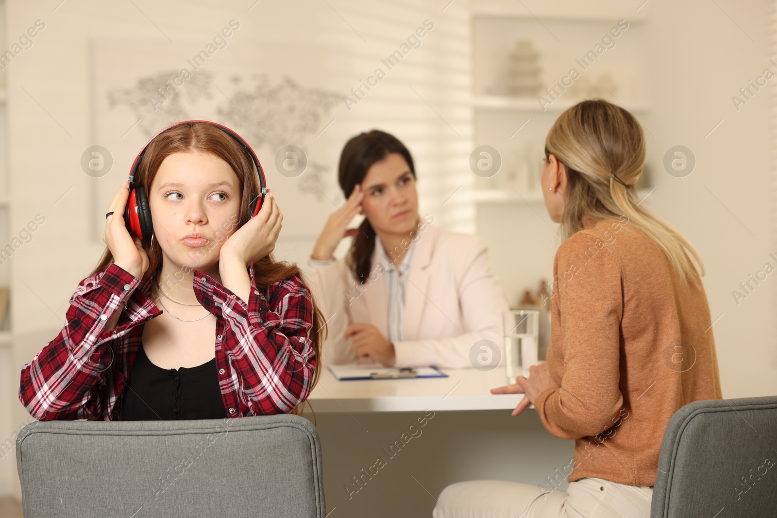 Photo of Annoyed teenage girl listening to music instead of working with psychologist in office, selective focus. Mother and daughter difficult relationship