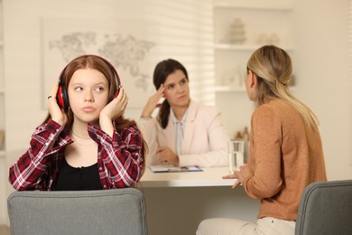 Annoyed teenage girl listening to music instead of working with psychologist in office, selective focus. Mother and daughter difficult relationship