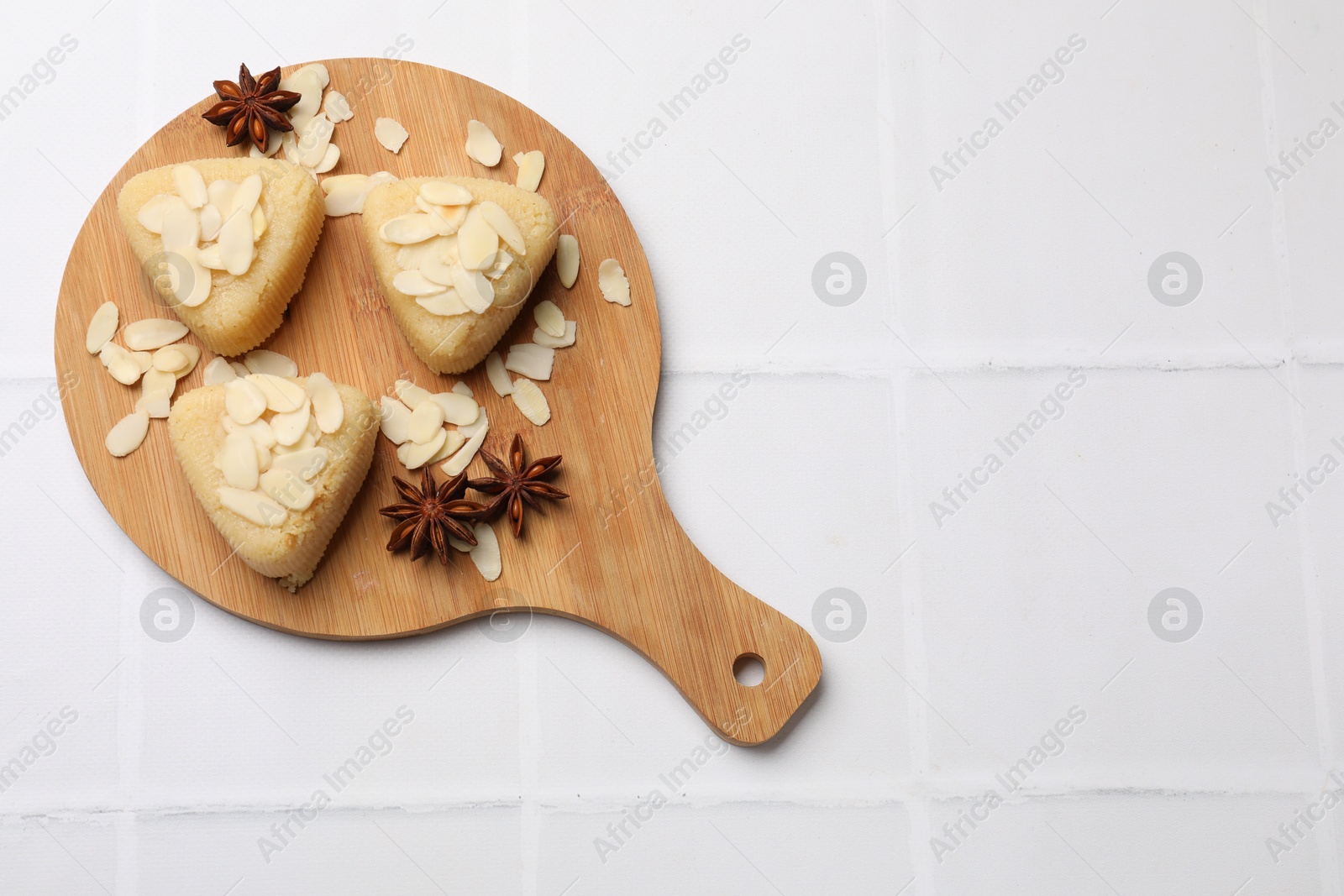 Photo of Pieces of delicious sweet semolina halva with almond flakes on white tiled table, top view. Space for text