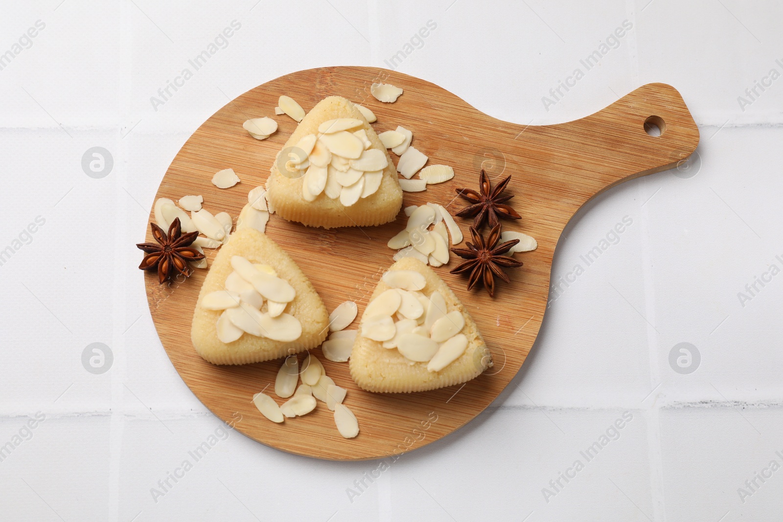 Photo of Pieces of delicious sweet semolina halva with almond flakes on white tiled table, top view