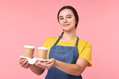 Photo of Girl in apron with takeaway paper cups of coffee on pink background. Work for teenagers