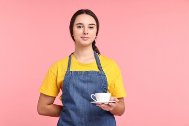 Photo of Girl in apron with cup of coffee on pink background. Work for teenagers