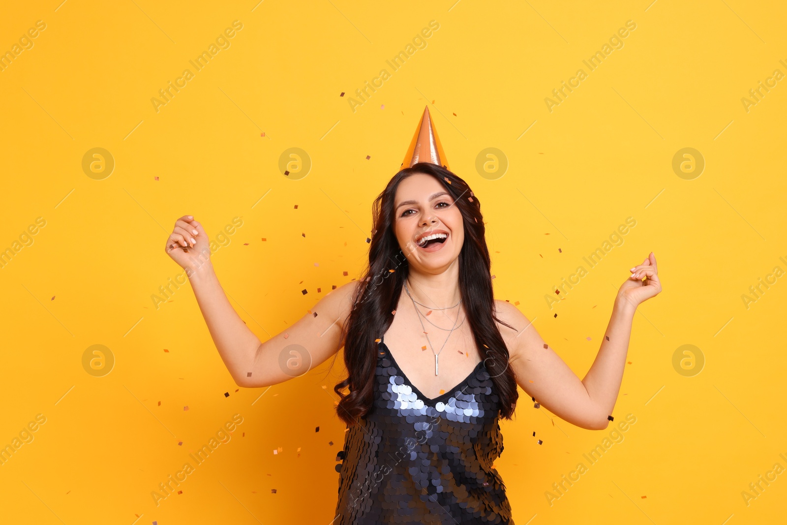 Photo of Happy woman in conical paper hat under falling confetti on orange background. Surprise party