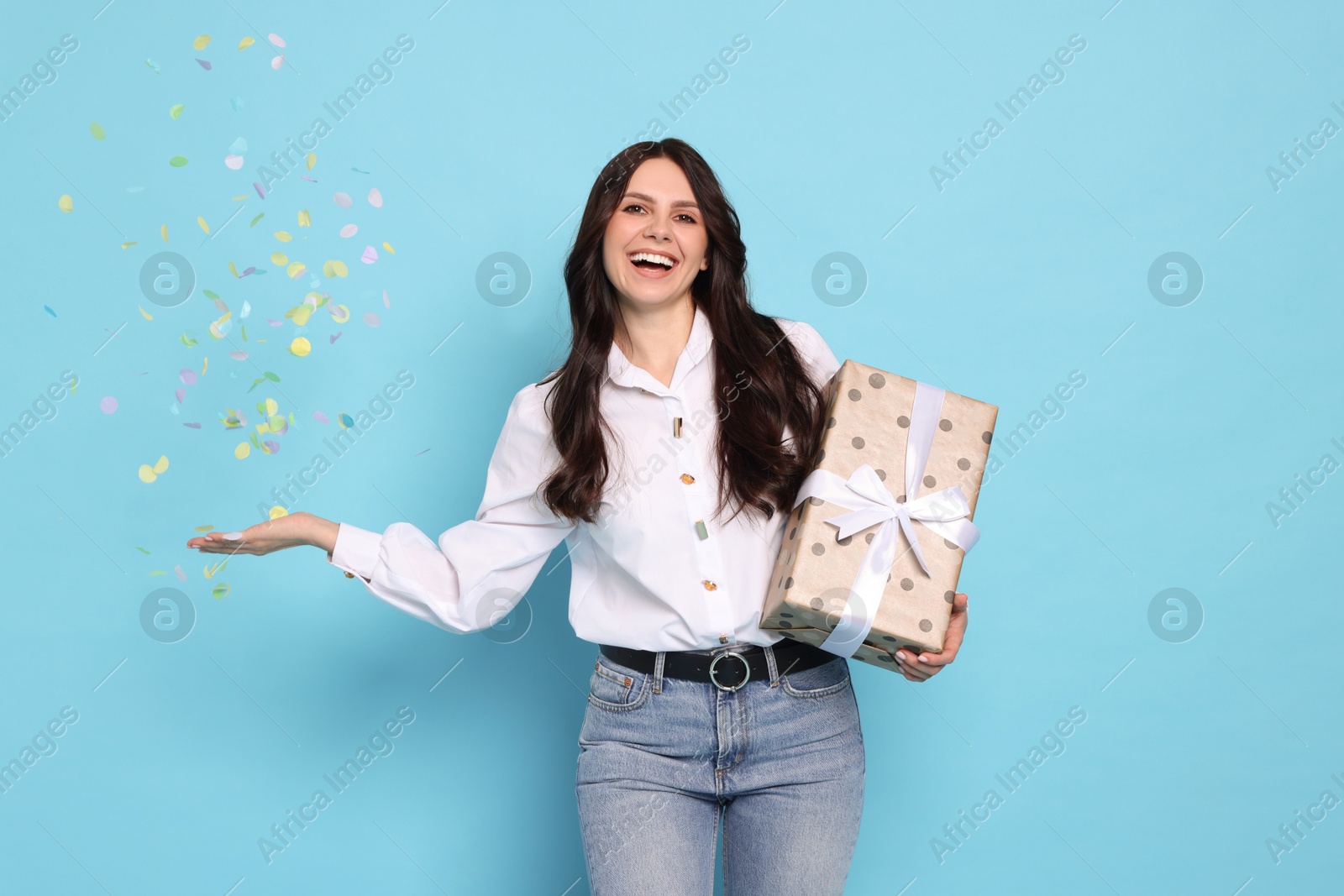 Photo of Happy woman with gift box and flying confetti on light blue background. Surprise party