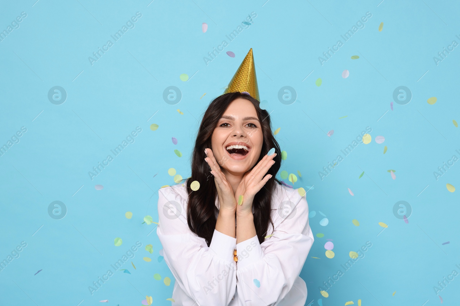 Photo of Happy woman in conical paper hat under falling confetti on light blue background. Surprise party