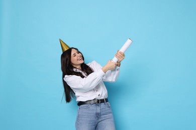 Happy woman in conical paper hat blowing up confetti popper on light blue background. Surprise party