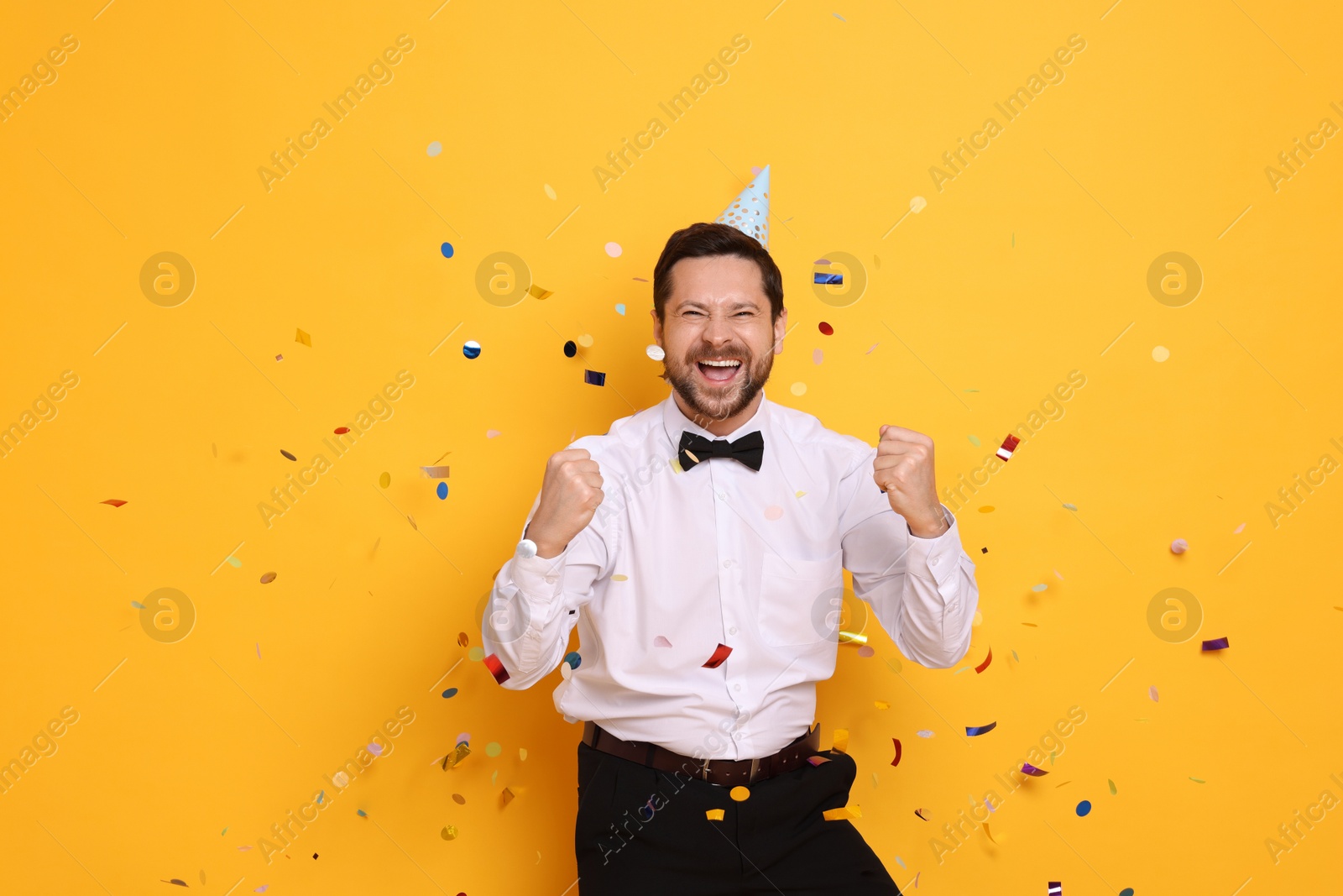 Photo of Happy man in conical paper hat and flying confetti on orange background. Surprise party