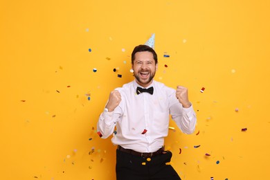 Photo of Happy man in conical paper hat and flying confetti on orange background. Surprise party