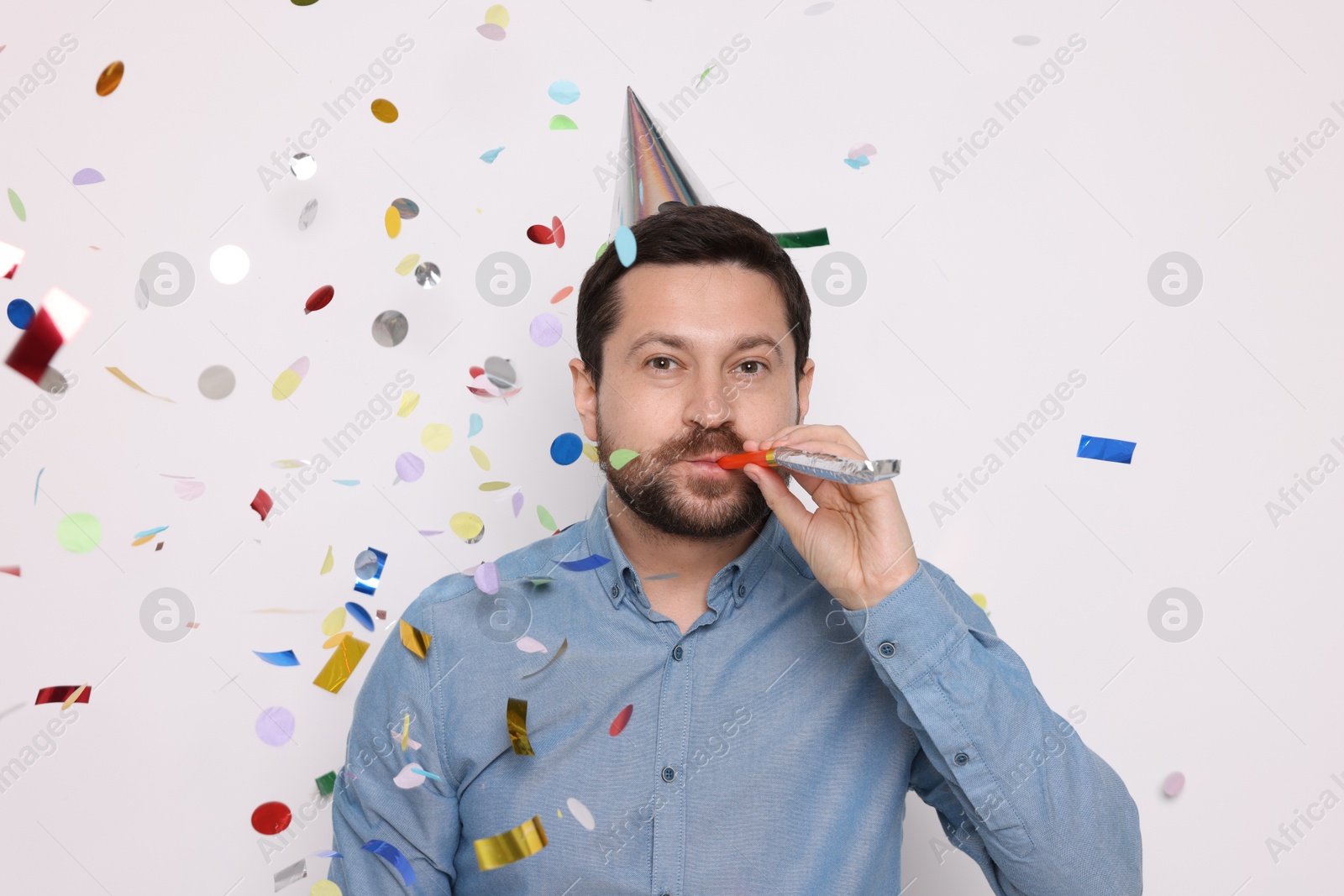 Photo of Man in conical paper hat with blower and falling confetti on white background. Surprise party