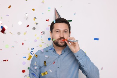 Photo of Man in conical paper hat with blower and falling confetti on white background. Surprise party