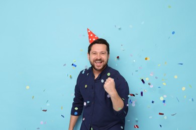 Photo of Happy man in conical paper hat under falling confetti on light blue background. Surprise party