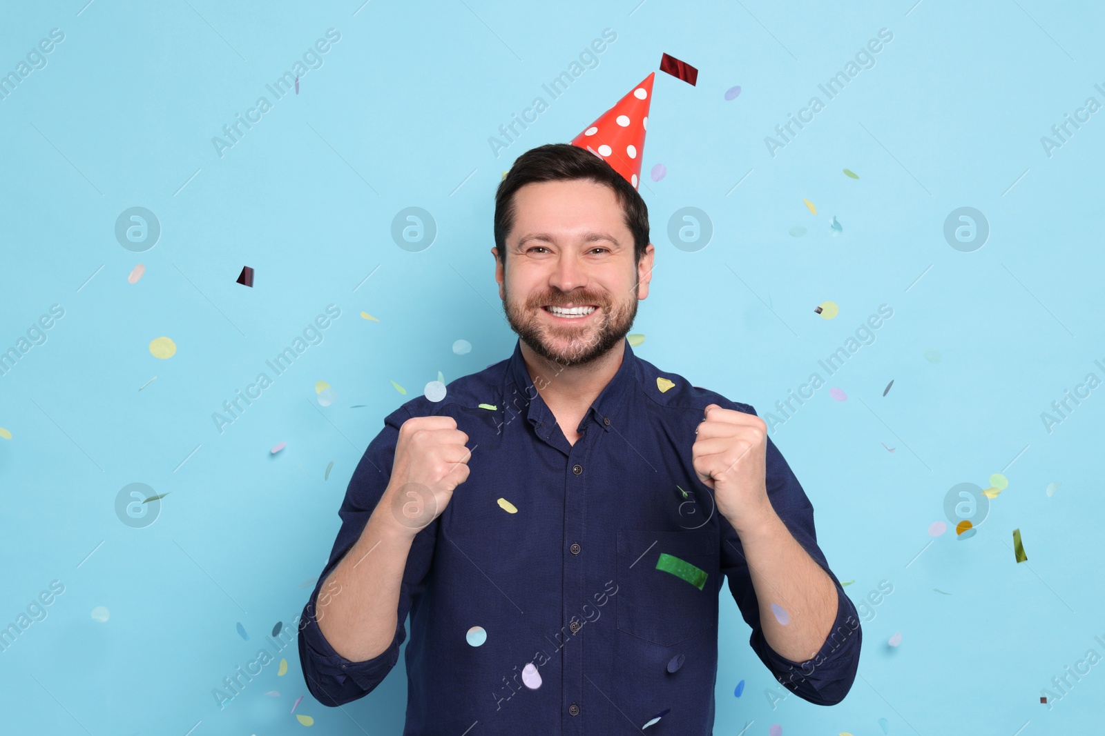 Photo of Happy man in conical paper hat under falling confetti on light blue background. Surprise party