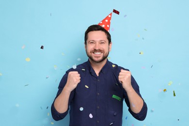 Happy man in conical paper hat under falling confetti on light blue background. Surprise party