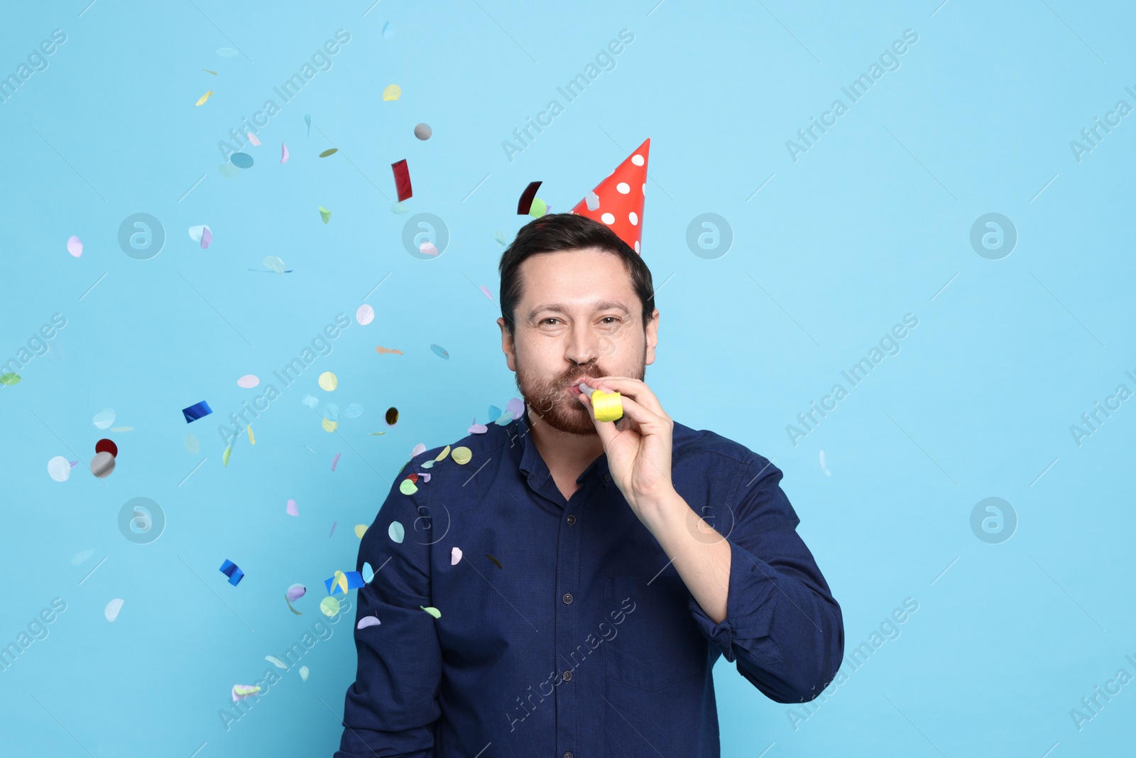 Photo of Man in conical paper hat with blower and flying confetti on light blue background. Surprise party