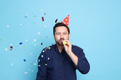 Man in conical paper hat with blower and flying confetti on light blue background. Surprise party