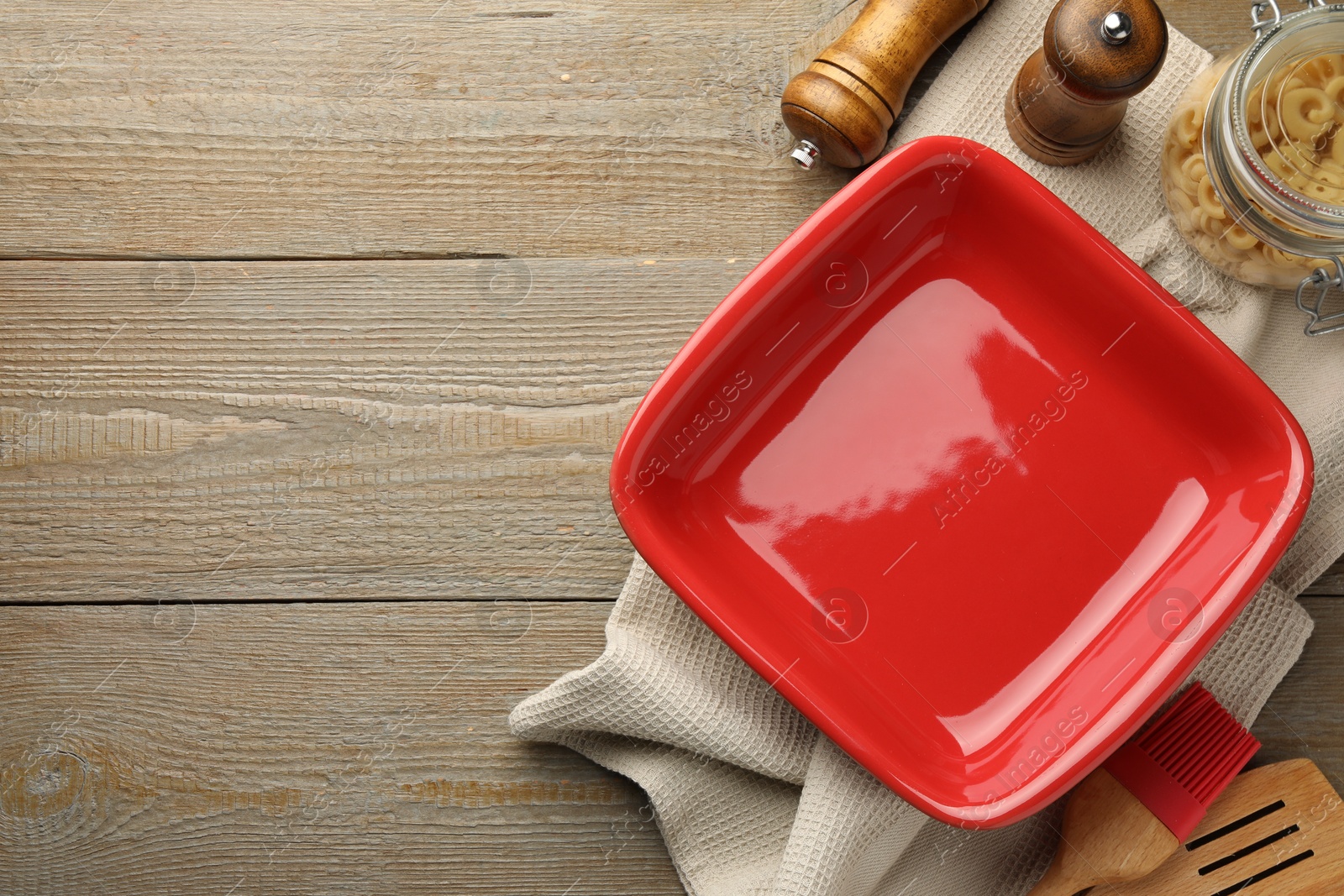 Photo of Ceramic casserole, raw pasta, shakers, brush and slotted turner on wooden table, flat lay. Space for text