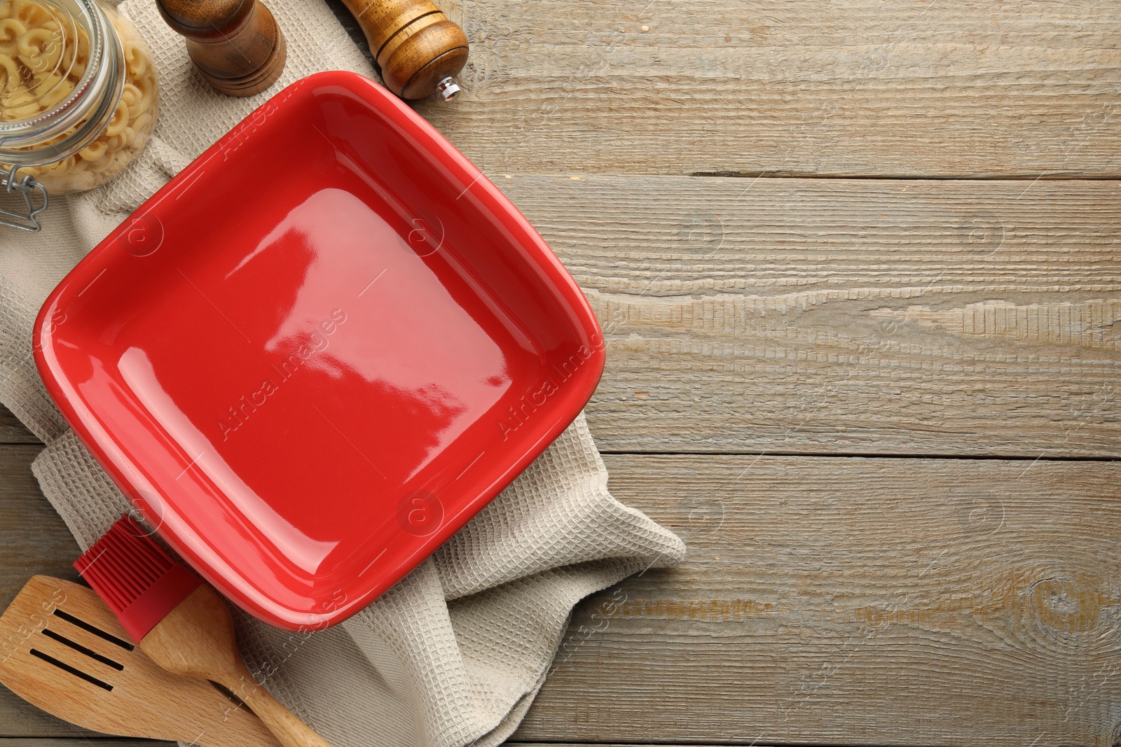 Photo of Ceramic casserole, raw pasta, shakers, brush and slotted turner on wooden table, flat lay. Space for text