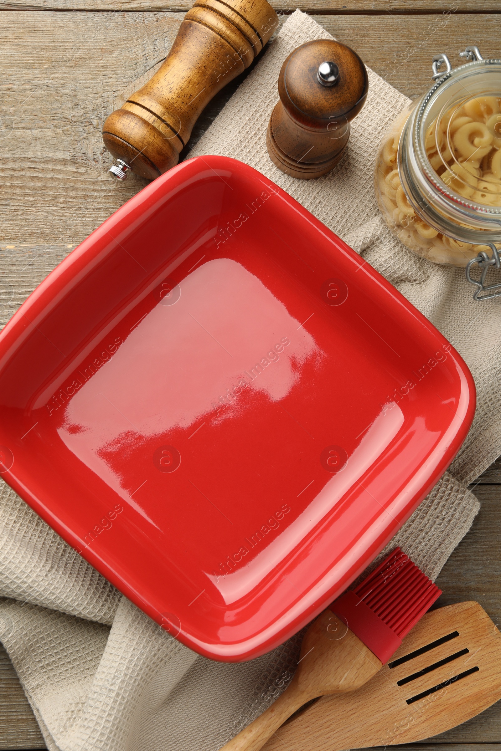 Photo of Ceramic casserole, raw pasta, shakers, brush and slotted turner on wooden table, flat lay