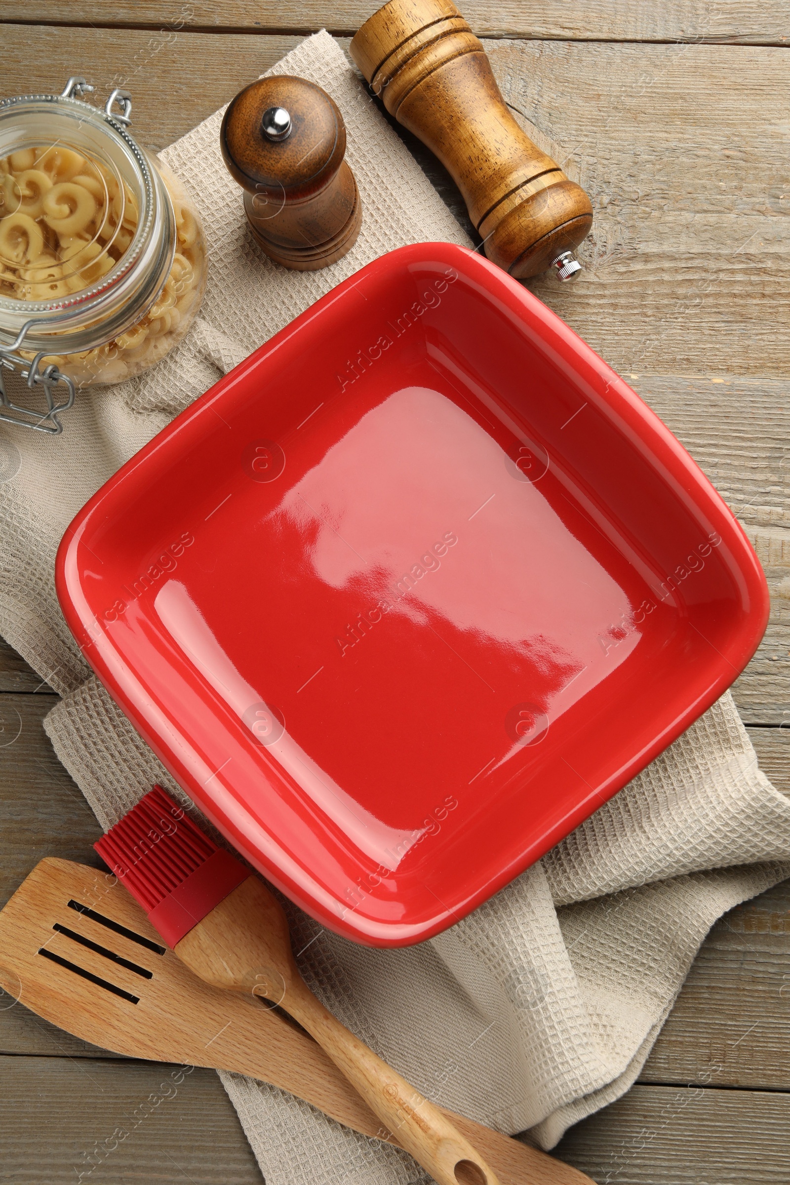 Photo of Ceramic casserole, raw pasta, shakers, brush and slotted turner on wooden table, flat lay