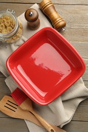 Photo of Ceramic casserole, raw pasta, shakers, brush and slotted turner on wooden table, flat lay