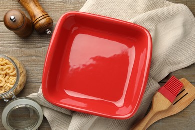 Photo of Ceramic casserole, raw pasta, shakers, brush and slotted turner on wooden table, flat lay