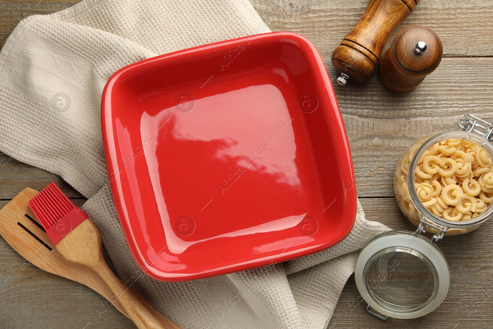 Photo of Ceramic casserole, raw pasta, shakers, brush and slotted turner on wooden table, flat lay