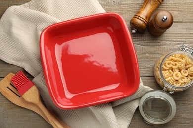 Photo of Ceramic casserole, raw pasta, shakers, brush and slotted turner on wooden table, flat lay