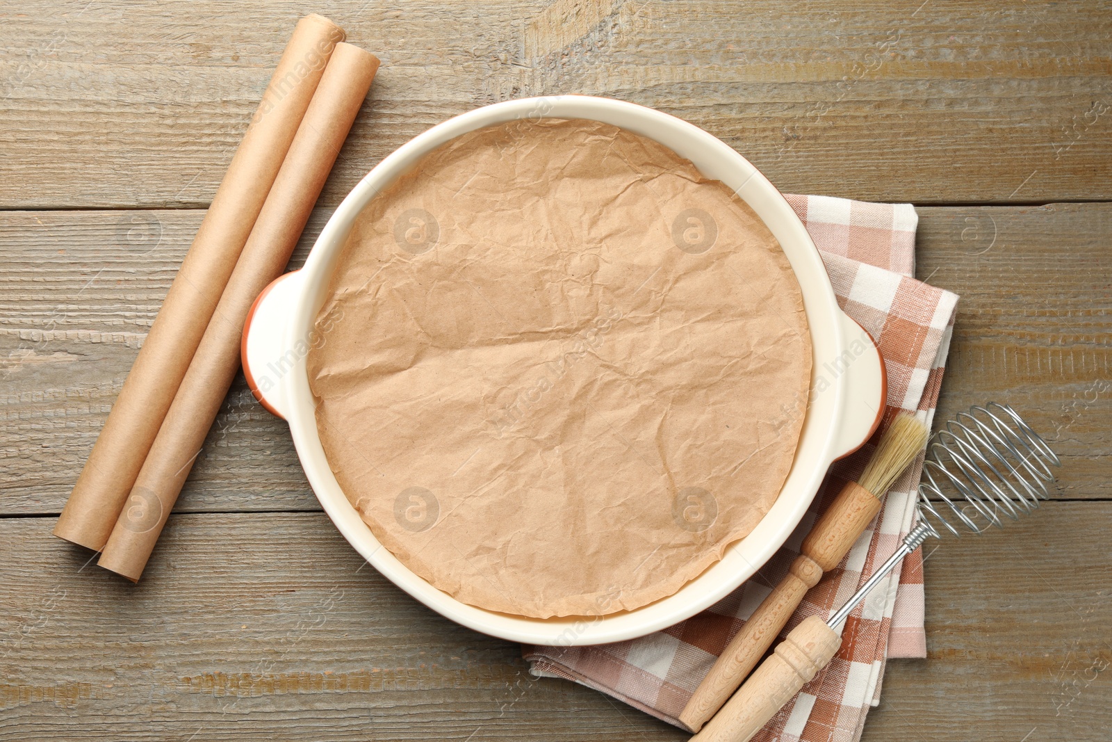 Photo of Ceramic casserole, baking parchment paper, brush and whisk on wooden table, flat lay