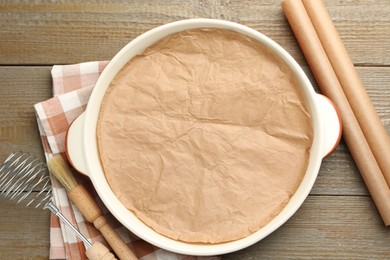 Photo of Ceramic casserole, baking parchment paper, brush and whisk on wooden table, flat lay