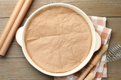 Photo of Ceramic casserole, baking parchment paper, brush and whisk on wooden table, flat lay