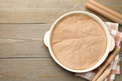 Photo of Ceramic casserole, baking parchment paper, brush and whisk on wooden table, flat lay. Space for text