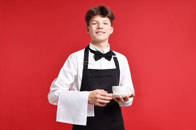 Photo of Teenage boy with cloth and cup of drink working as waiter on red background