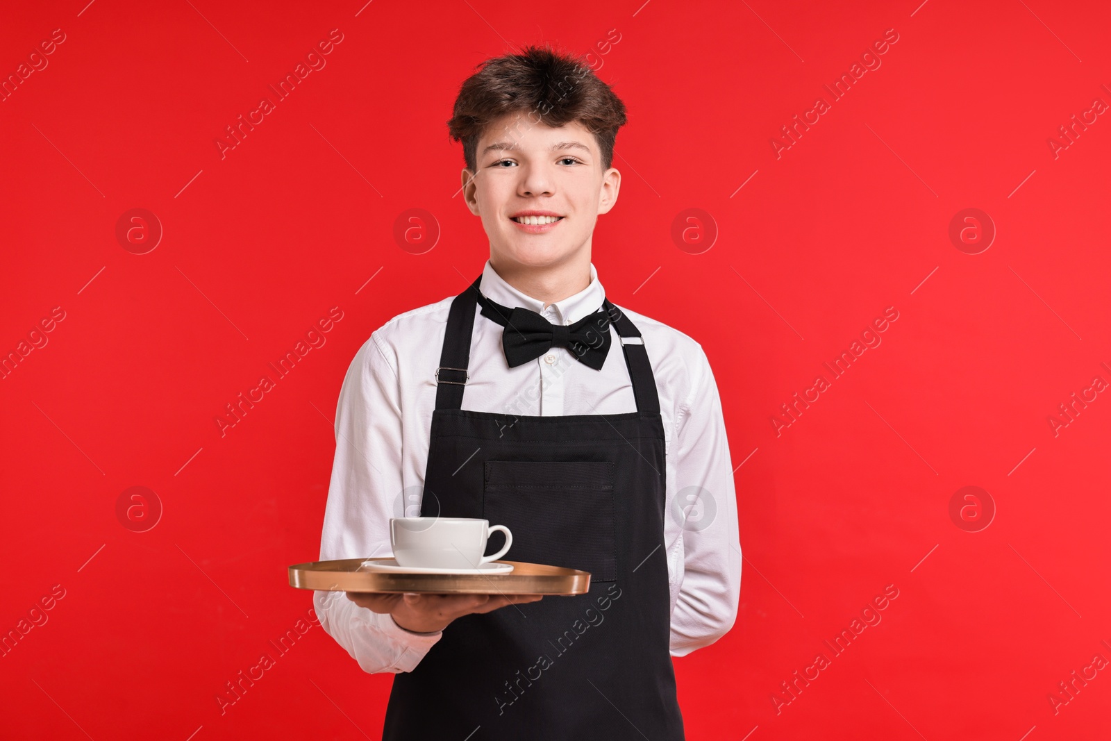 Photo of Teenage boy with cup of drink working as waiter on red background