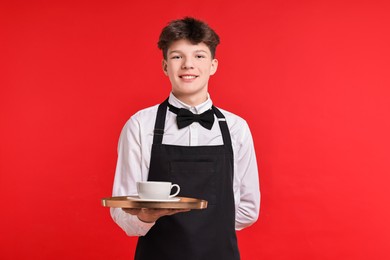 Photo of Teenage boy with cup of drink working as waiter on red background