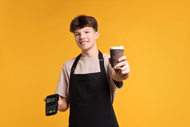 Photo of Teenage boy with paper cup and payment terminal working as cashier on orange background