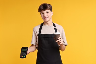 Photo of Teenage boy with paper cup and payment terminal working as cashier on orange background