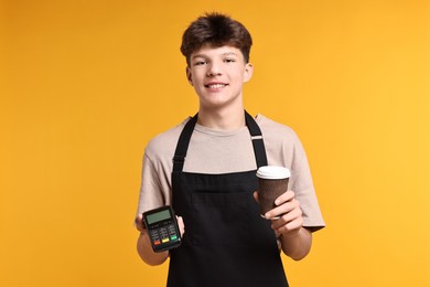 Photo of Teenage boy with paper cup and payment terminal working as cashier on orange background