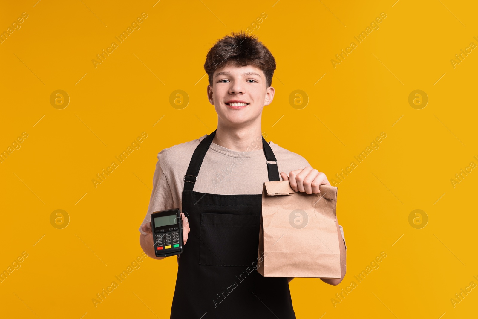 Photo of Teenage boy with paper bag and payment terminal working as cashier on orange background