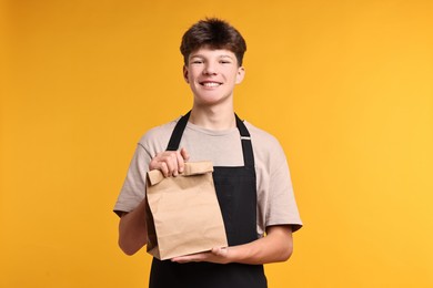 Teenage boy with paper bag working as cashier on orange background