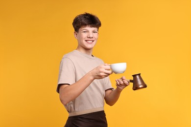 Photo of Teenage boy with cezve pot and cup working as barista on orange background